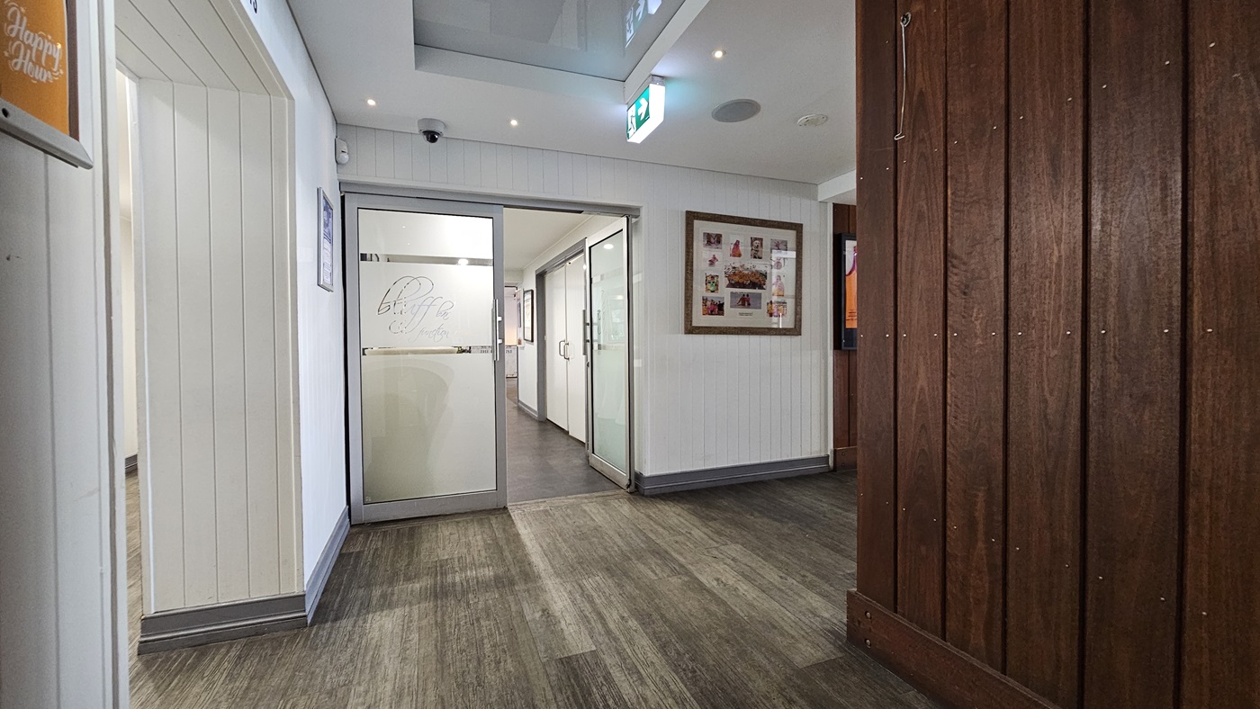 On the left, there is a white panelled wall with a small framed picture and a doorway leading to a hallway. In the middle, there is a glass door with the word "Bluff" etched on it, opening into another room with white walls and dark flooring. In the background, there are more glass doors and a lit exit sign. On the right, there is a wooden panelled wall with a large framed collage of photos. The floor is a mix of dark and light wood, and the ceiling is white with recessed lighting. On the left, there is a white panelled wall with a small framed picture and a doorway leading to a hallway. In the middle, there is a glass door with the word "Bluff" etched on it, opening into another room with white walls and dark flooring. In the background, there are more glass doors and a lit exit sign. On the right, there is a wooden panelled wall with a large framed collage of photos. The floor is a mix of dark and light wood, and the ceiling is white with recessed lighting.