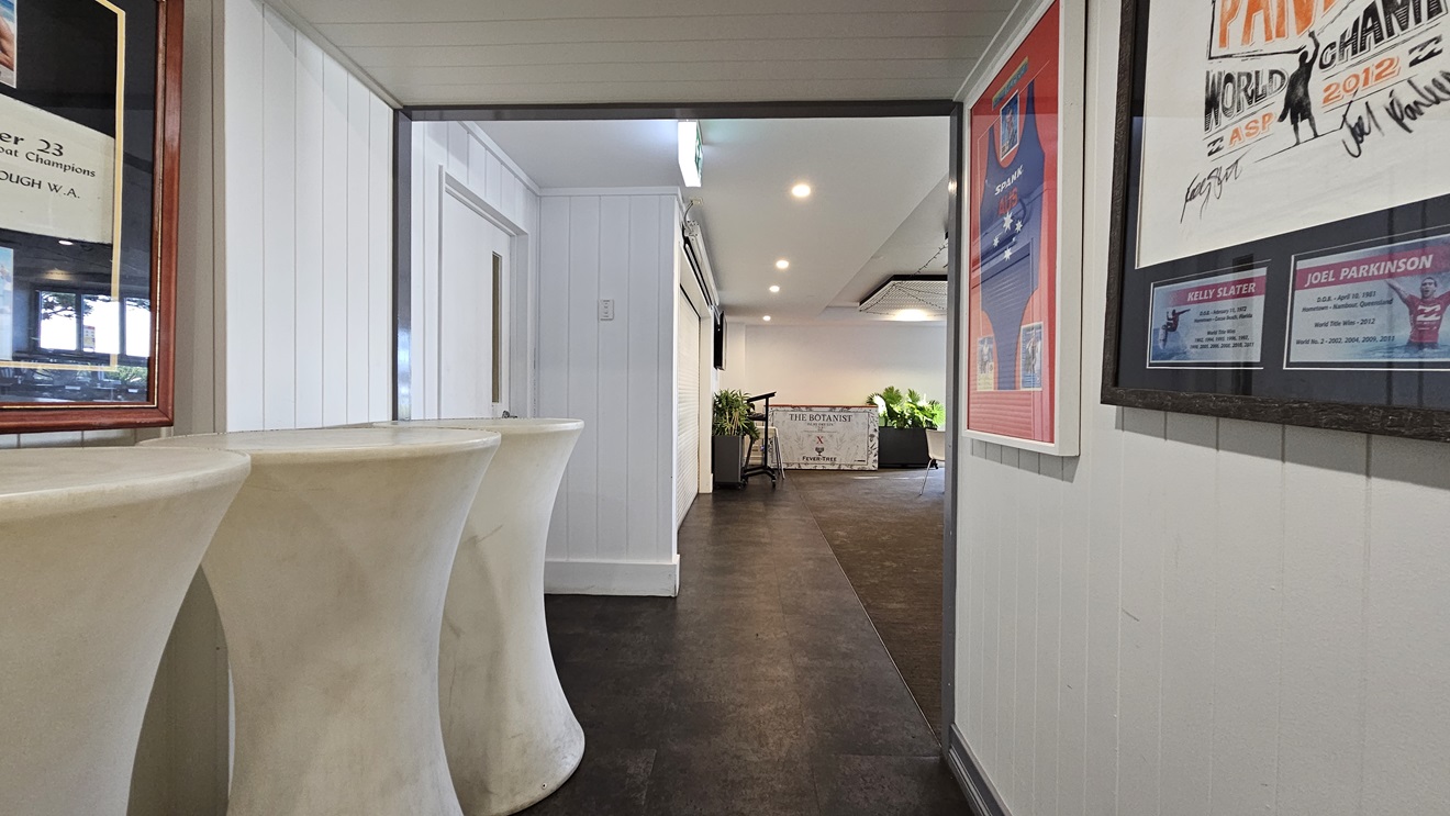On the left, there are tall, white pedestal tables and a white panelled wall with framed pictures. In the middle, there is a hallway with dark flooring leading to a well-lit area with plants and a white table. In the background, there is a white wall with a door and more plants. On the right, there is a white panelled wall with framed posters and a bulletin board. The ceiling is white with recessed lighting. On the left, there are tall, white pedestal tables and a white panelled wall with framed pictures. In the middle, there is a hallway with dark flooring leading to a well-lit area with plants and a white table. In the background, there is a white wall with a door and more plants. On the right, there is a white panelled wall with framed posters and a bulletin board. The ceiling is white with recessed lighting.