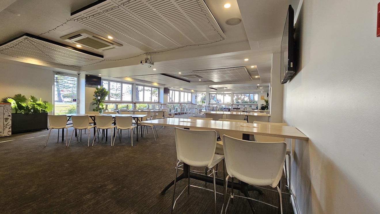 On the left, there are several tables with white chairs and potted plants near the windows. In the middle, there are more tables and chairs arranged in rows, with large windows in the background letting in natural light. On the right, there is a white wall with a mounted TV and a long table with white chairs. The floor is covered in dark carpet, and the ceiling has white panels with recessed lighting and air conditioning units. On the left, there are several tables with white chairs and potted plants near the windows. In the middle, there are more tables and chairs arranged in rows, with large windows in the background letting in natural light. On the right, there is a white wall with a mounted TV and a long table with white chairs. The floor is covered in dark carpet, and the ceiling has white panels with recessed lighting and air conditioning units.