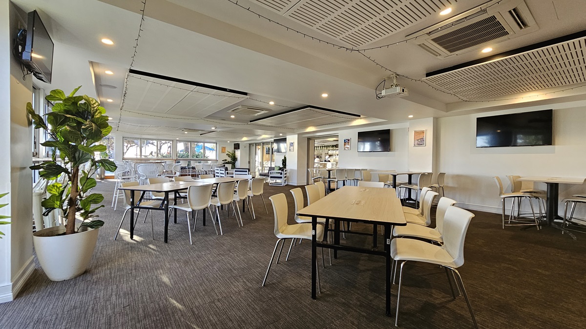 On the left, there are several tables with white chairs and a large potted plant near the windows. In the middle, there are more tables and chairs arranged in rows, with large windows in the background letting in natural light. On the right, there are two wall-mounted TVs above a long table with white chairs. The floor is covered in dark carpet, and the ceiling has white panels with recessed lighting and air conditioning units. On the left, there are several tables with white chairs and a large potted plant near the windows. In the middle, there are more tables and chairs arranged in rows, with large windows in the background letting in natural light. On the right, there are two wall-mounted TVs above a long table with white chairs. The floor is covered in dark carpet, and the ceiling has white panels with recessed lighting and air conditioning units.