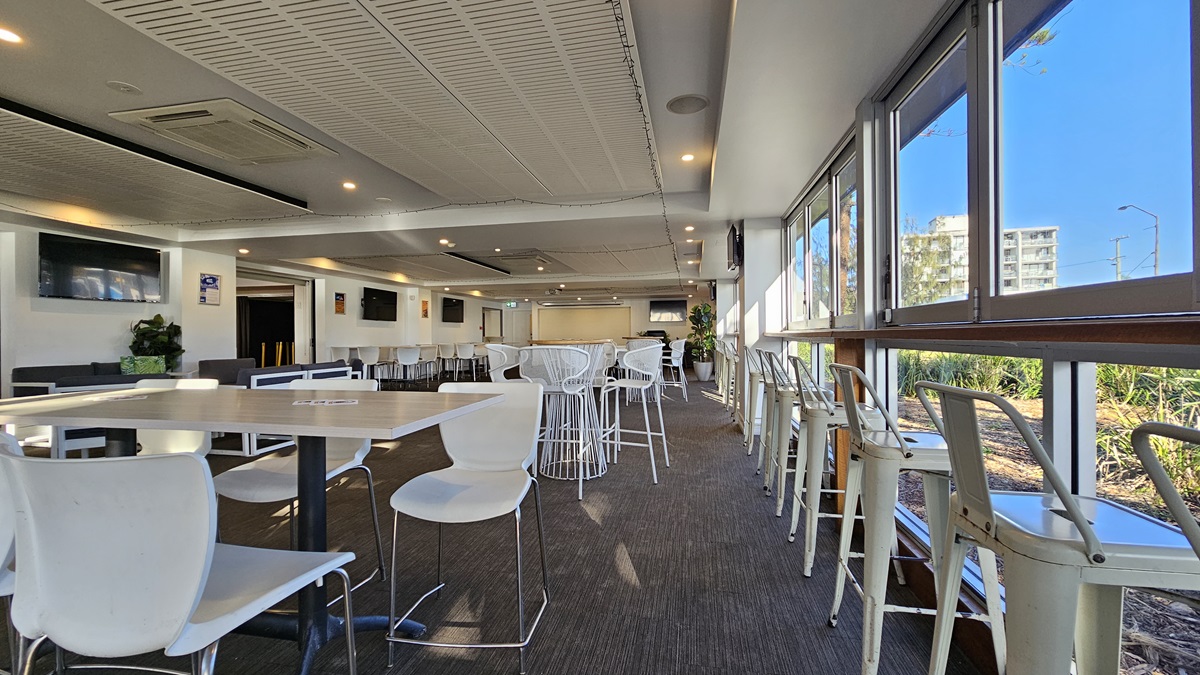 On the left, there are white tables with white chairs and a wall-mounted TV above a potted plant. In the middle, there are more white tables and chairs, with a long counter and bar stools in the background. On the right, there are large windows with high stools and tables along the window line, offering a view of greenery outside. The floor is covered in dark carpet, and the ceiling has white panels with recessed lighting and air conditioning units. On the left, there are white tables with white chairs and a wall-mounted TV above a potted plant. In the middle, there are more white tables and chairs, with a long counter and bar stools in the background. On the right, there are large windows with high stools and tables along the window line, offering a view of greenery outside. The floor is covered in dark carpet, and the ceiling has white panels with recessed lighting and air conditioning units.