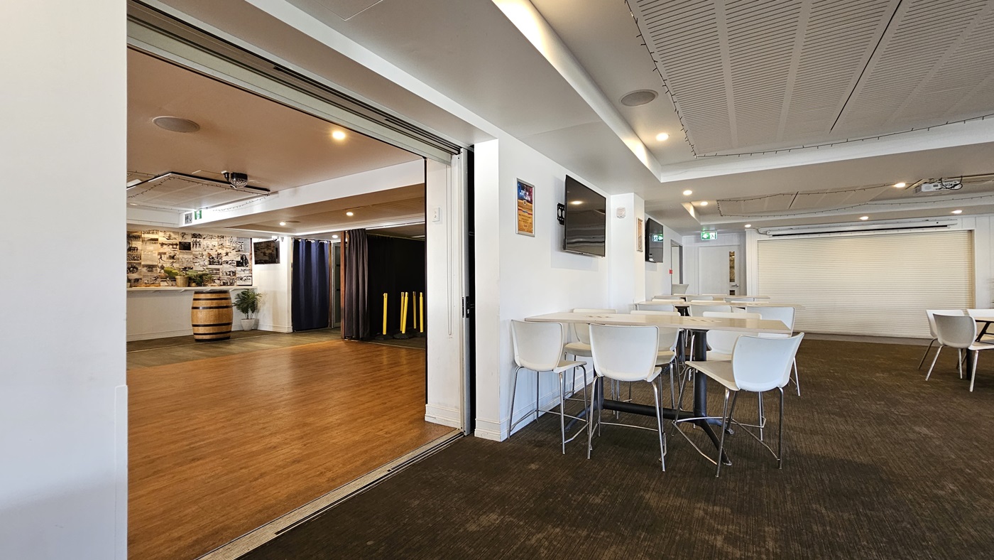 On the left, there is an open area with wooden flooring, a large barrel, and a wall covered in photos. In the middle, there is a partition wall separating the wooden floor area from the carpeted area. In the foreground, there are white tables and chairs on dark carpet. On the right, there is a wall-mounted TV above a white table and chairs, and a large roller door in the background. The ceiling has white panels with recessed lighting and air conditioning vents. On the left, there is an open area with wooden flooring, a large barrel, and a wall covered in photos. In the middle, there is a partition wall separating the wooden floor area from the carpeted area. In the foreground, there are white tables and chairs on dark carpet. On the right, there is a wall-mounted TV above a white table and chairs, and a large roller door in the background. The ceiling has white panels with recessed lighting and air conditioning vents.