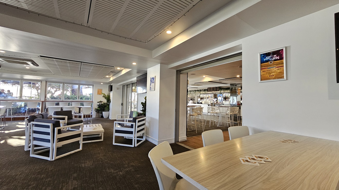 On the left, there are white-framed lounge chairs with dark cushions around low tables, and large windows with a view of trees. In the middle, there is a partition wall with a doorway leading to a bar area in the background, featuring high stools and a counter. On the right, there is a wooden table with white chairs and a framed poster on the wall. The floor is covered in dark carpet, and the ceiling has white panels with recessed lighting and air conditioning vents. On the left, there are white-framed lounge chairs with dark cushions around low tables, and large windows with a view of trees. In the middle, there is a partition wall with a doorway leading to a bar area in the background, featuring high stools and a counter. On the right, there is a wooden table with white chairs and a framed poster on the wall. The floor is covered in dark carpet, and the ceiling has white panels with recessed lighting and air conditioning vents.