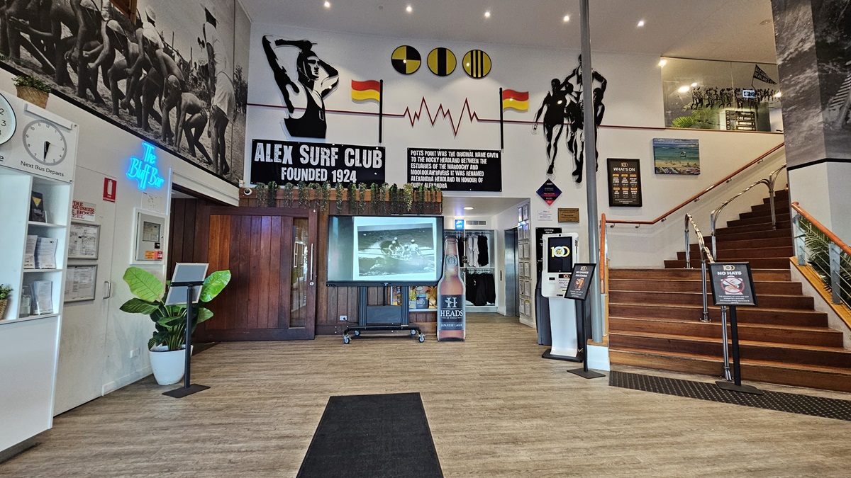 On the left, there is a wooden door with a "Bar" sign above it, a clock, and a potted plant. In the middle, a large screen on a stand displays an image, with surf club memorabilia and wall art above it. On the right, a staircase with wooden steps and a metal handrail leads to an upper level. The background includes more surf-themed decorations and a glass railing. The floor is wooden with a black mat in the centre, and the ceiling has recessed lighting. On the left, there is a wooden door with a "Bar" sign above it, a clock, and a potted plant. In the middle, a large screen on a stand displays an image, with surf club memorabilia and wall art above it. On the right, a staircase with wooden steps and a metal handrail leads to an upper level. The background includes more surf-themed decorations and a glass railing. The floor is wooden with a black mat in the centre, and the ceiling has recessed lighting.