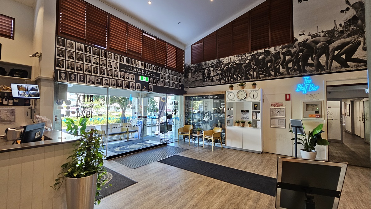 On the left, there is a reception desk with a plant and a computer monitor displaying security footage. In the middle, there are two yellow chairs with cushions and a small round table between them, in front of a glass display case filled with trophies and historical photos. On the right, there is a wooden door with a "Bar" sign above it, a potted plant, and a hallway leading to other rooms. The floor is a mix of wooden and carpeted areas, and the ceiling has recessed lighting and wooden blinds. On the left, there is a reception desk with a plant and a computer monitor displaying security footage. In the middle, there are two yellow chairs with cushions and a small round table between them, in front of a glass display case filled with trophies and historical photos. On the right, there is a wooden door with a "Bar" sign above it, a potted plant, and a hallway leading to other rooms. The floor is a mix of wooden and carpeted areas, and the ceiling has recessed lighting and wooden blinds.
