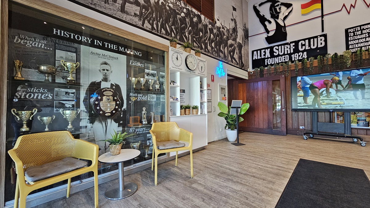 On the left, there are two yellow chairs with cushions and a small round table between them, in front of a glass display case filled with trophies and historical photos. In the middle, there is a wall with clocks and a potted plant. On the right, a large screen displays a beach scene, next to a wooden door with a "Bar" sign above it. The background features surf club memorabilia and wall art. The floor is wooden with a black mat in the centre, and the ceiling has recessed lighting. On the left, there are two yellow chairs with cushions and a small round table between them, in front of a glass display case filled with trophies and historical photos. In the middle, there is a wall with clocks and a potted plant. On the right, a large screen displays a beach scene, next to a wooden door with a "Bar" sign above it. The background features surf club memorabilia and wall art. The floor is wooden with a black mat in the centre, and the ceiling has recessed lighting.