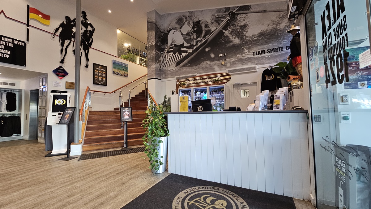 On the left, there is a staircase with wooden steps and a metal handrail, leading to an upper level. In the middle, there is a wall with surf-themed artwork and a large black-and-white mural of surfers above a reception counter. On the right, the reception counter has a plant, merchandise, and a display case with drinks. The floor is wooden with a black mat in front of the counter, and the ceiling has recessed lighting and surf club flags. On the left, there is a staircase with wooden steps and a metal handrail, leading to an upper level. In the middle, there is a wall with surf-themed artwork and a large black-and-white mural of surfers above a reception counter. On the right, the reception counter has a plant, merchandise, and a display case with drinks. The floor is wooden with a black mat in front of the counter, and the ceiling has recessed lighting and surf club flags.
