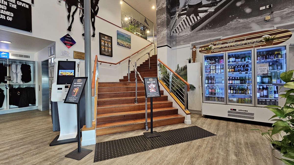 On the left, there is a kiosk with a touchscreen and a sign above it, next to a display of black clothing. In the middle, a wooden staircase with a metal handrail leads to an upper level, with surf-themed artwork and a large black-and-white mural of surfers on the wall above. On the right, there is a large refrigerated display case filled with drinks, and a potted plant beside it. The floor is wooden with a black mat at the base of the stairs, and the ceiling has recessed lighting. On the left, there is a kiosk with a touchscreen and a sign above it, next to a display of black clothing. In the middle, a wooden staircase with a metal handrail leads to an upper level, with surf-themed artwork and a large black-and-white mural of surfers on the wall above. On the right, there is a large refrigerated display case filled with drinks, and a potted plant beside it. The floor is wooden with a black mat at the base of the stairs, and the ceiling has recessed lighting.