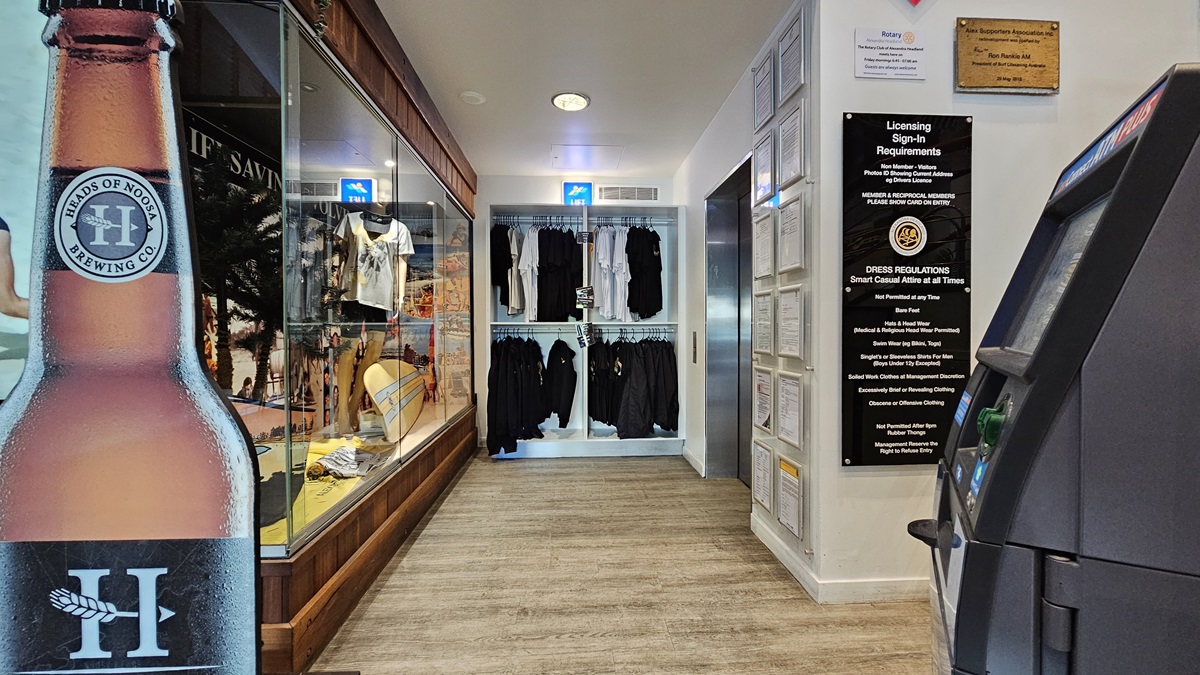 On the left, there is a large poster of a beer bottle next to a glass display case containing various items, including clothing and surfboards. In the middle, there are shelves with neatly arranged black clothing. On the right, there is an ATM machine and a wall with licensing signs and requirements. The floor is wooden, and the ceiling has recessed lighting and a smoke detector. On the left, there is a large poster of a beer bottle next to a glass display case containing various items, including clothing and surfboards. In the middle, there are shelves with neatly arranged black clothing. On the right, there is an ATM machine and a wall with licensing signs and requirements. The floor is wooden, and the ceiling has recessed lighting and a smoke detector.