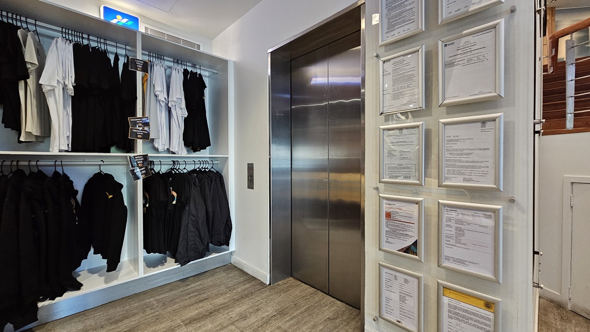 On the left, there are shelves with neatly arranged black and white clothing on hangers. In the middle, there is a stainless steel lift with closed doors and a control panel on the wall beside it. On the right, there is a wall with multiple framed documents and certificates. The floor is wooden, and the ceiling has recessed lighting and an air vent. On the left, there are shelves with neatly arranged black and white clothing on hangers. In the middle, there is a stainless steel lift with closed doors and a control panel on the wall beside it. On the right, there is a wall with multiple framed documents and certificates. The floor is wooden, and the ceiling has recessed lighting and an air vent.