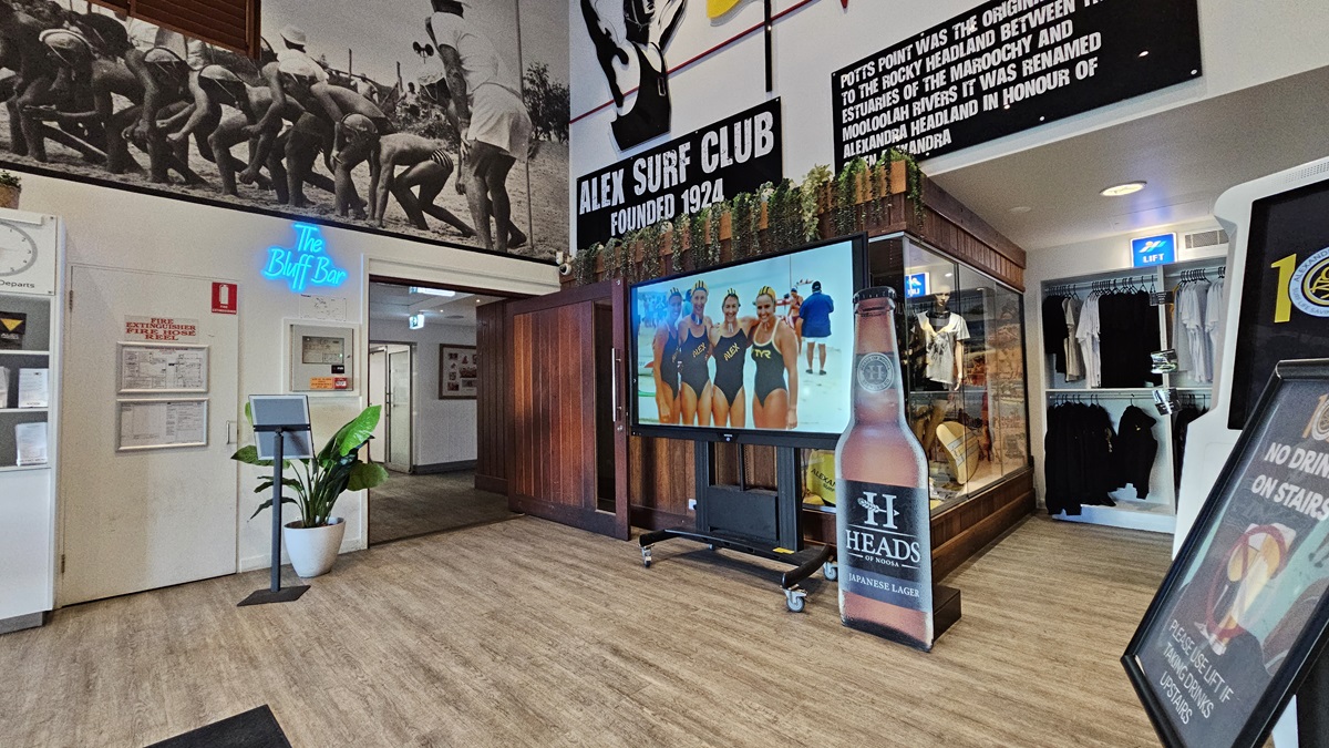 On the left, there is a white wall with a blue neon sign reading "The Buff Bar" above a small counter with a plant beside it. In the middle, there is a large screen on a stand displaying an image of people in swimsuits, with a surf club mural above it. On the right, there is a glass display case with surf club memorabilia and a blackboard with writing. The floor is wooden, and the ceiling has recessed lighting and surf club flags. On the left, there is a white wall with a blue neon sign reading "The Buff Bar" above a small counter with a plant beside it. In the middle, there is a large screen on a stand displaying an image of people in swimsuits, with a surf club mural above it. On the right, there is a glass display case with surf club memorabilia and a blackboard with writing. The floor is wooden, and the ceiling has recessed lighting and surf club flags.