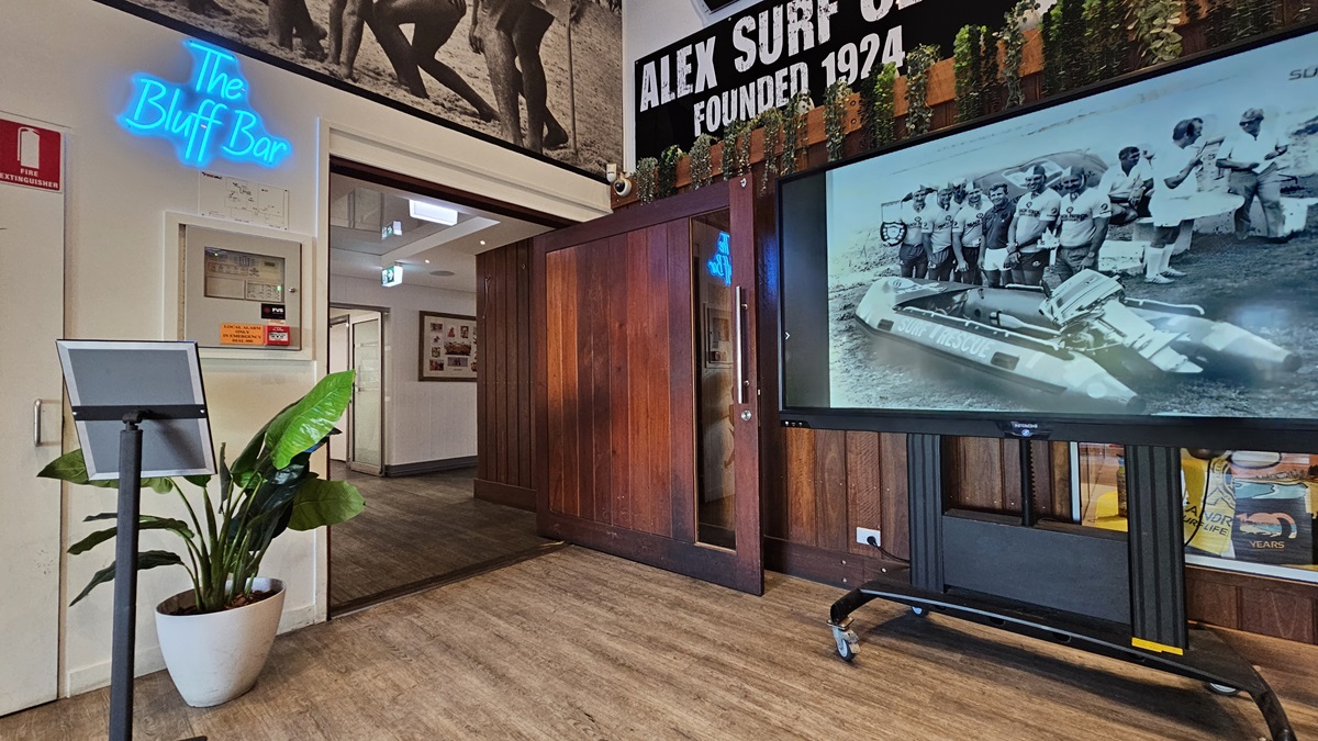 On the left, there is a white wall with a blue neon sign reading "The Bluff Bar" above a small counter with a plant beside it. In the middle, there is a wooden doorway leading to a hallway with framed photos on the walls. On the right, there is a large screen on a stand displaying a black-and-white image of people with a boat, and a glass display case with surf club memorabilia below it. The floor is wooden, and the ceiling has recessed lighting and surf club flags. On the left, there is a white wall with a blue neon sign reading "The Bluff Bar" above a small counter with a plant beside it. In the middle, there is a wooden doorway leading to a hallway with framed photos on the walls. On the right, there is a large screen on a stand displaying a black-and-white image of people with a boat, and a glass display case with surf club memorabilia below it. The floor is wooden, and the ceiling has recessed lighting and surf club flags.