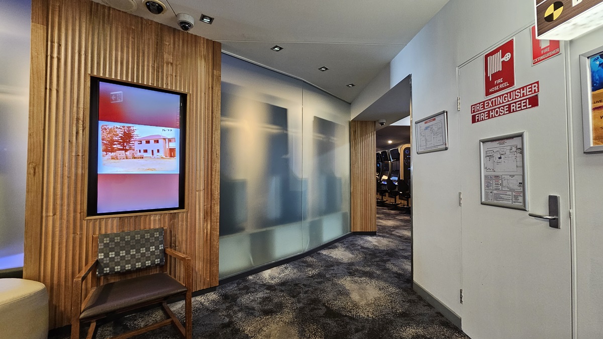On the left, there is a wooden wall with vertical slats and a digital screen displaying an image of a building. Below the screen, there is a wooden chair with a patterned cushion. On the right, there is a white wall with fire safety signs, a fire extinguisher, and a door. In the middle, there is a frosted glass partition leading to a hallway with more chairs. The floor is carpeted with a dark, mottled pattern, and the ceiling has white panels with recessed lighting. On the left, there is a wooden wall with vertical slats and a digital screen displaying an image of a building. Below the screen, there is a wooden chair with a patterned cushion. On the right, there is a white wall with fire safety signs, a fire extinguisher, and a door. In the middle, there is a frosted glass partition leading to a hallway with more chairs. The floor is carpeted with a dark, mottled pattern, and the ceiling has white panels with recessed lighting.