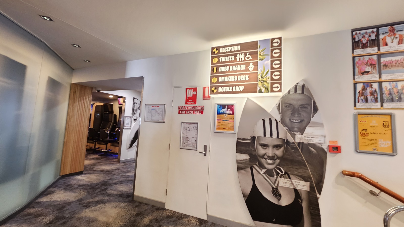 On the left, there is a wall with black and white photos of people and a wooden floor leading to a hallway. On the right, there is an ATM machine with a small trash bin next to it. In the middle, there is a decorative partition with circular cutouts. The floor is a mix of wooden and dark carpeted areas, and the ceiling has recessed lighting. On the left, there is a wall with black and white photos of people and a wooden floor leading to a hallway. On the right, there is an ATM machine with a small trash bin next to it. In the middle, there is a decorative partition with circular cutouts. The floor is a mix of wooden and dark carpeted areas, and the ceiling has recessed lighting.