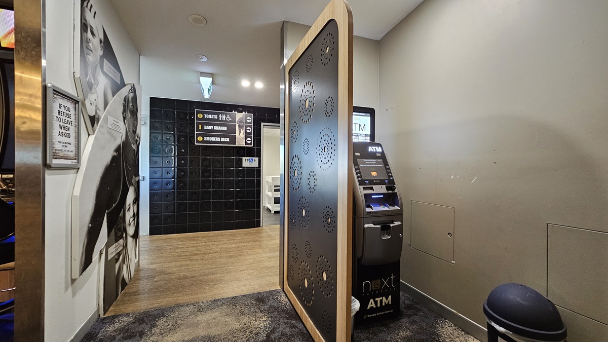 On the left, there is a wall with black and white photos of people and a wooden floor leading to a hallway. On the right, there is an ATM machine with a small trash bin next to it. In the middle, there is a decorative partition with circular cutouts. The floor is a mix of wooden and dark carpeted areas, and the ceiling has recessed lighting. On the left, there is a wall with black and white photos of people and a wooden floor leading to a hallway. On the right, there is an ATM machine with a small trash bin next to it. In the middle, there is a decorative partition with circular cutouts. The floor is a mix of wooden and dark carpeted areas, and the ceiling has recessed lighting.