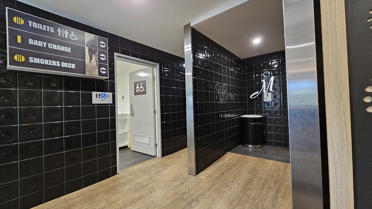 On the left, there is a sign indicating toilets, baby change, and smokers deck, above a doorway leading to a baby change room. On the right, there is a black tiled wall with a silver bin and a sign with the letter "M" for men's toilets. In the middle, there is a corridor with black tiled walls and a wooden floor. The ceiling is grey with recessed lighting. On the left, there is a sign indicating toilets, baby change, and smokers deck, above a doorway leading to a baby change room. On the right, there is a black tiled wall with a silver bin and a sign with the letter "M" for men's toilets. In the middle, there is a corridor with black tiled walls and a wooden floor. The ceiling is grey with recessed lighting.