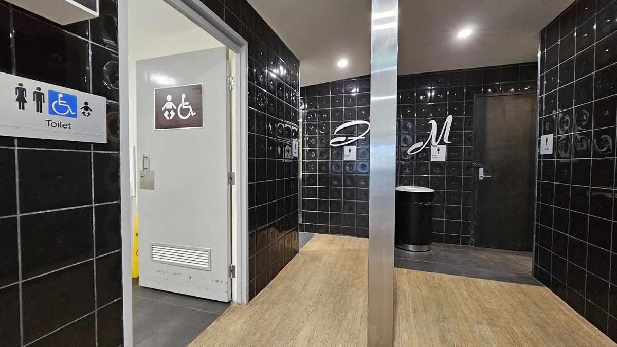 On the left, there is a doorway with a sign indicating accessible toilets, baby change, and a smokers' deck, leading to a baby change room with a white door. On the right, there is a black tiled wall with a silver bin and a sign with the letter "M" for men's toilets next to a black door. In the middle, there is a corridor with black tiled walls and a wooden floor. The ceiling is grey with recessed lighting. On the left, there is a doorway with a sign indicating accessible toilets, baby change, and a smokers' deck, leading to a baby change room with a white door. On the right, there is a black tiled wall with a silver bin and a sign with the letter "M" for men's toilets next to a black door. In the middle, there is a corridor with black tiled walls and a wooden floor. The ceiling is grey with recessed lighting.