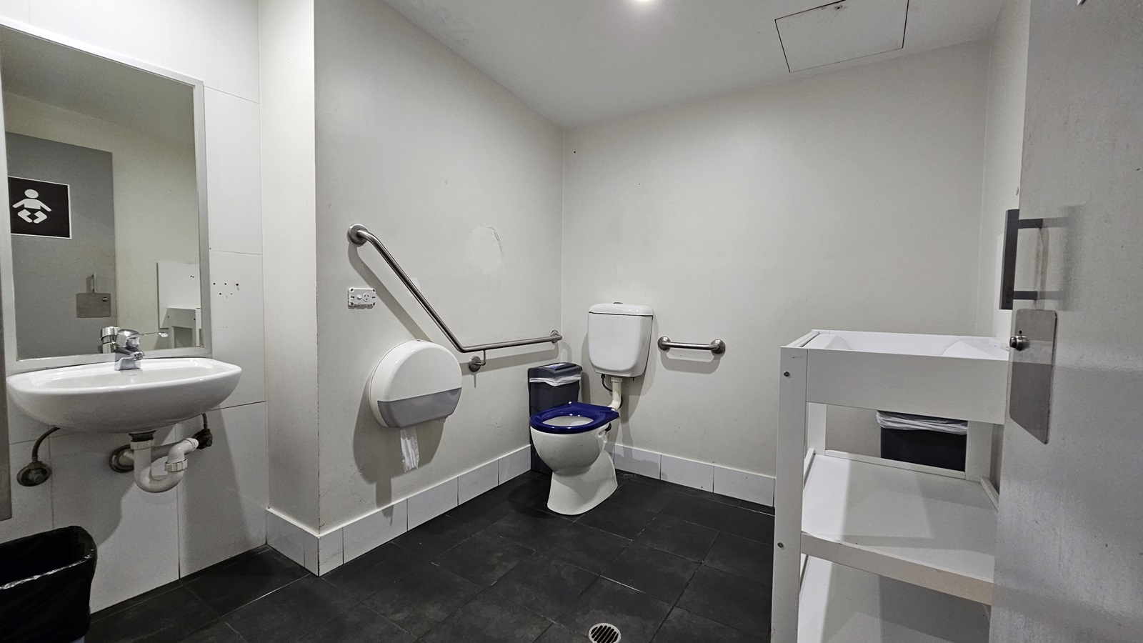 On the left, there is a white sink with a mirror above it and a black bin below. Next to the sink, there is a wall-mounted baby changing station. On the right, there is a white shelving unit with a bin on the bottom shelf. In the middle, there is a toilet with a blue seat, a grab rail on the wall, and a toilet paper dispenser. The floor is covered with black tiles, and the ceiling is white with recessed lighting. On the left, there is a white sink with a mirror above it and a black bin below. Next to the sink, there is a wall-mounted baby changing station. On the right, there is a white shelving unit with a bin on the bottom shelf. In the middle, there is a toilet with a blue seat, a grab rail on the wall, and a toilet paper dispenser. The floor is covered with black tiles, and the ceiling is white with recessed lighting.