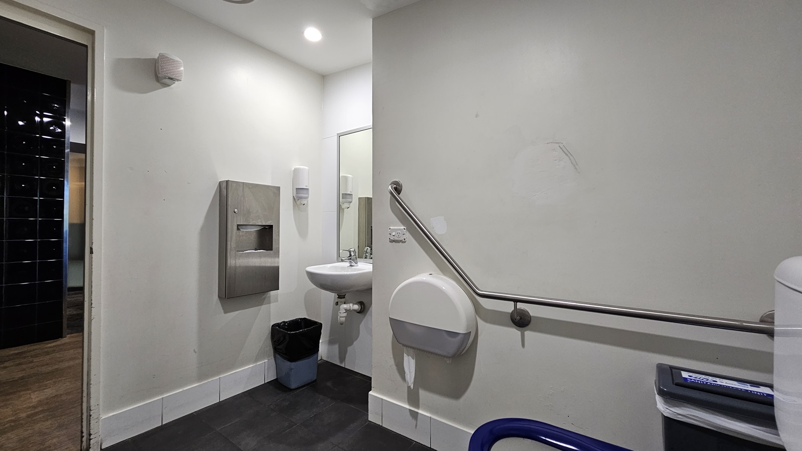 On the left, there is a doorway leading to a hallway with dark tiles. Next to the doorway, there is a wall-mounted paper towel dispenser above a black bin. Further right, there is a white sink with a mirror above it and soap dispensers on either side. On the right, there is a grab rail mounted on the wall, a wall-mounted baby changing station, and a toilet with a blue seat. The floor is covered with dark tiles, and the ceiling is white with recessed lighting. On the left, there is a doorway leading to a hallway with dark tiles. Next to the doorway, there is a wall-mounted paper towel dispenser above a black bin. Further right, there is a white sink with a mirror above it and soap dispensers on either side. On the right, there is a grab rail mounted on the wall, a wall-mounted baby changing station, and a toilet with a blue seat. The floor is covered with dark tiles, and the ceiling is white with recessed lighting.