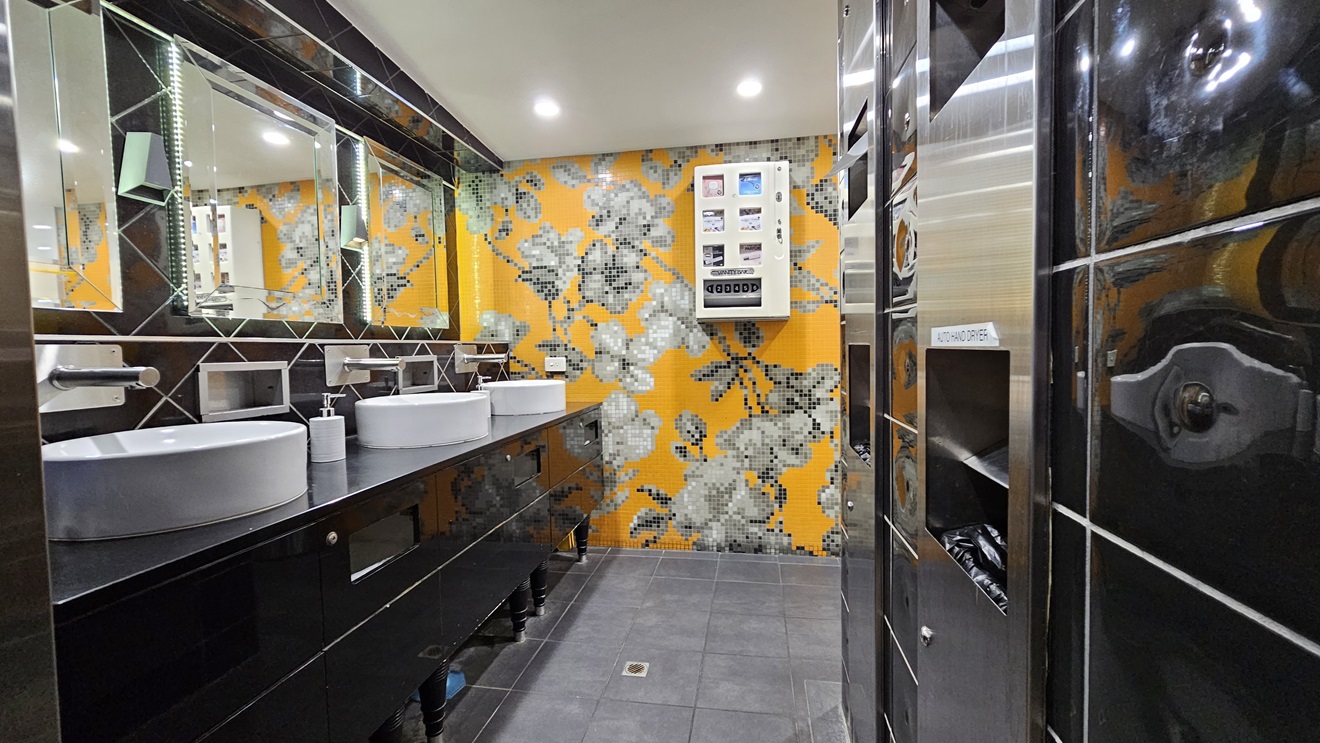 On the left, there are two white sinks with mirrors above them, set on a black countertop with soap dispensers. Below the countertop, there are storage shelves. On the right, there is a black tiled wall with a silver hand dryer and a vending machine. In the background, the wall is covered with orange and grey floral mosaic tiles. The floor is covered with dark grey tiles, and the ceiling is white with recessed lighting. On the left, there are two white sinks with mirrors above them, set on a black countertop with soap dispensers. Below the countertop, there are storage shelves. On the right, there is a black tiled wall with a silver hand dryer and a vending machine. In the background, the wall is covered with orange and grey floral mosaic tiles. The floor is covered with dark grey tiles, and the ceiling is white with recessed lighting.