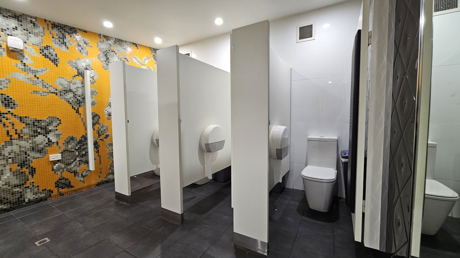 On the left, there is a wall covered with orange and grey floral mosaic tiles. In the middle, there are three white toilet cubicles with partial walls and doors, each containing a toilet and a large toilet paper dispenser. On the right, there is a white tiled wall with a full-length mirror. The floor is covered with dark grey tiles, and the ceiling is white with recessed lighting. On the left, there is a wall covered with orange and grey floral mosaic tiles. In the middle, there are three white toilet cubicles with partial walls and doors, each containing a toilet and a large toilet paper dispenser. On the right, there is a white tiled wall with a full-length mirror. The floor is covered with dark grey tiles, and the ceiling is white with recessed lighting.
