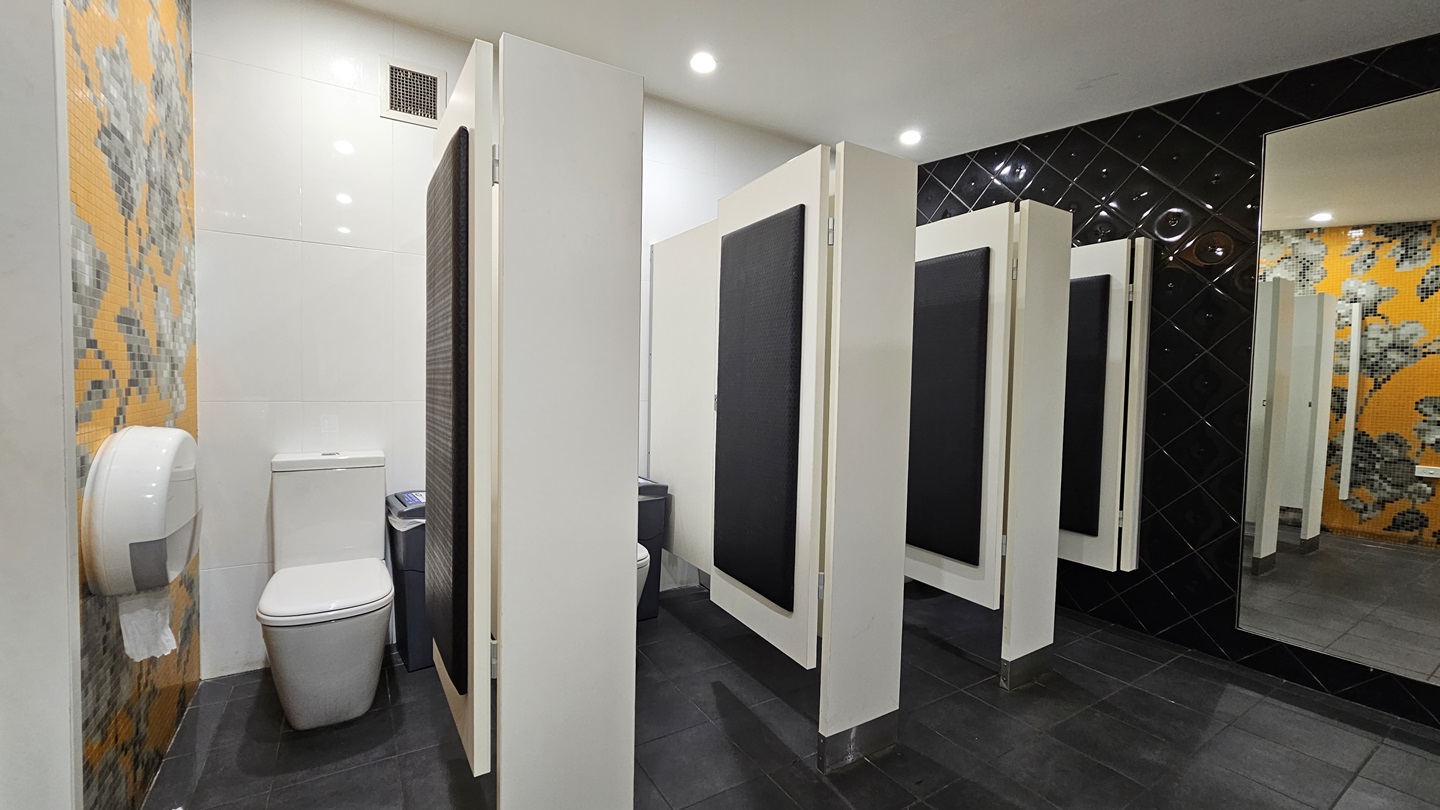 On the left, there is a toilet with a large toilet paper dispenser mounted on a wall covered with orange and grey floral mosaic tiles. In the middle, there are three white toilet cubicles with black doors, each containing a toilet. On the right, there is a black tiled wall with a full-length mirror. The floor is covered with dark grey tiles, and the ceiling is white with recessed lighting. On the left, there is a toilet with a large toilet paper dispenser mounted on a wall covered with orange and grey floral mosaic tiles. In the middle, there are three white toilet cubicles with black doors, each containing a toilet. On the right, there is a black tiled wall with a full-length mirror. The floor is covered with dark grey tiles, and the ceiling is white with recessed lighting.