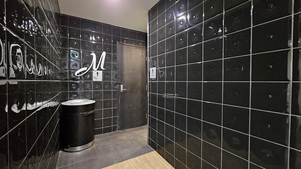 On the left, there is a black tiled wall with a silver bin and a sign with the letter "M" above a door. In the middle, there is a dark wooden door with a handle. On the right, there is another black tiled wall. The floor is covered with dark grey tiles, and the ceiling is white with recessed lighting. On the left, there is a black tiled wall with a silver bin and a sign with the letter "M" above a door. In the middle, there is a dark wooden door with a handle. On the right, there is another black tiled wall. The floor is covered with dark grey tiles, and the ceiling is white with recessed lighting.