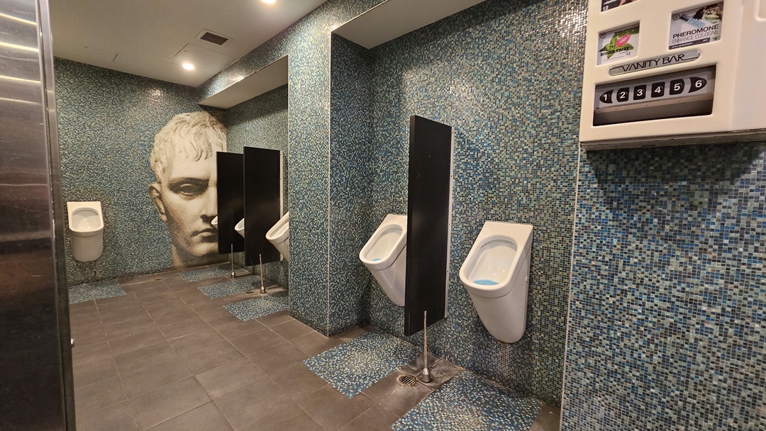 On the left, there is a wall with a large artistic face mural. In the middle, there are three white urinals separated by black dividers, mounted on a wall covered with blue mosaic tiles. On the right, there is a vending machine for toiletries attached to the same blue mosaic-tiled wall. The floor is covered with dark grey tiles, and the ceiling is white with recessed lighting. On the left, there is a wall with a large artistic face mural. In the middle, there are three white urinals separated by black dividers, mounted on a wall covered with blue mosaic tiles. On the right, there is a vending machine for toiletries attached to the same blue mosaic-tiled wall. The floor is covered with dark grey tiles, and the ceiling is white with recessed lighting.