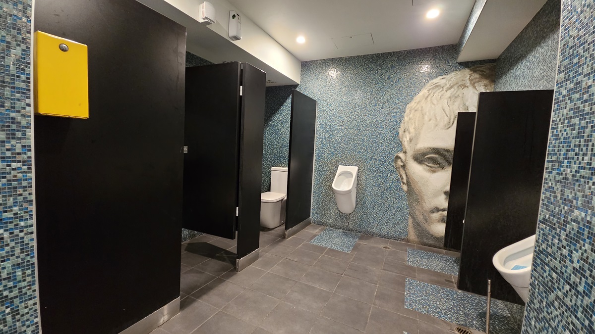 On the left, there are black cubicle doors with a yellow panel on one of them. In the middle, there is a white urinal mounted on a blue mosaic-tiled wall featuring a large mural of a face. On the right, there is a white sink with a blue mosaic-tiled backsplash. The floor is covered with dark grey tiles, and the ceiling is white with recessed lighting. On the left, there are black cubicle doors with a yellow panel on one of them. In the middle, there is a white urinal mounted on a blue mosaic-tiled wall featuring a large mural of a face. On the right, there is a white sink with a blue mosaic-tiled backsplash. The floor is covered with dark grey tiles, and the ceiling is white with recessed lighting.