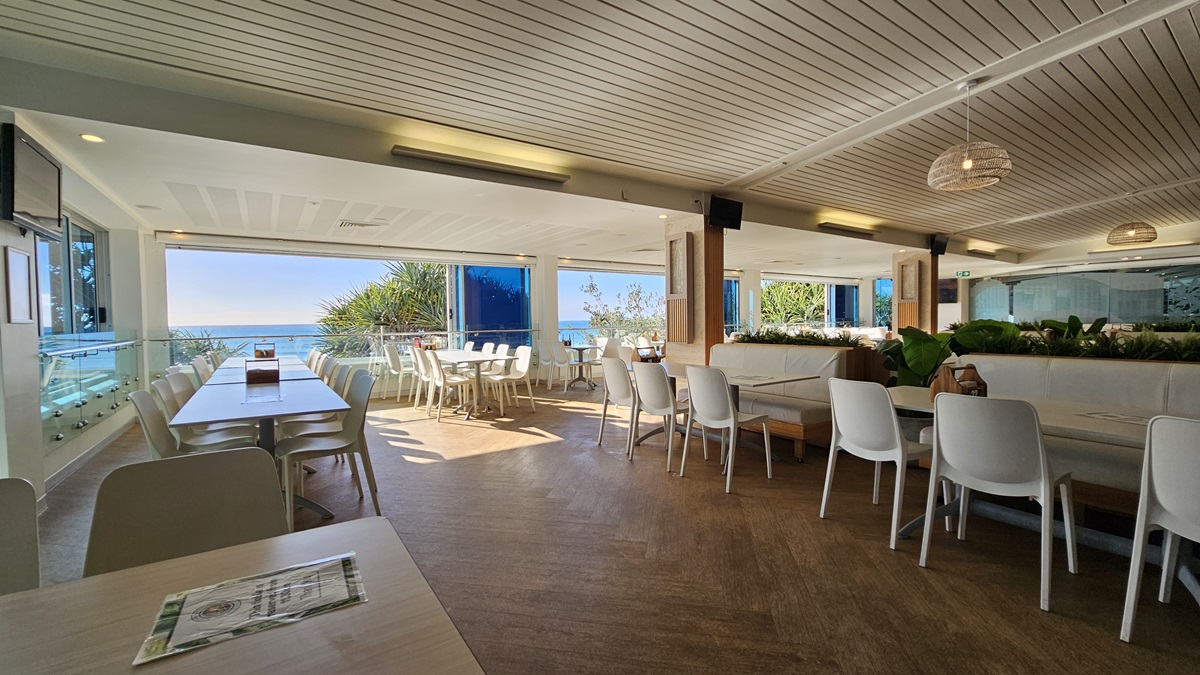 On the left, there are white tables and chairs near large windows with a view of the beach and trees. On the right, there are more white tables and chairs near floor-to-ceiling windows with blue-tinted glass. In the foreground, there are additional tables and chairs, with a wooden partition featuring potted plants on top. The floor is wooden, and the ceiling has white panels with recessed lighting, air conditioning vents, and hanging wicker lamps. On the left, there are white tables and chairs near large windows with a view of the beach and trees. On the right, there are more white tables and chairs near floor-to-ceiling windows with blue-tinted glass. In the foreground, there are additional tables and chairs, with a wooden partition featuring potted plants on top. The floor is wooden, and the ceiling has white panels with recessed lighting, air conditioning vents, and hanging wicker lamps.