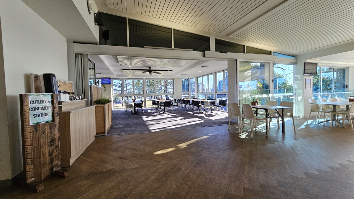 On the left, there is a wooden cutlery and condiment station next to a light wood counter. On the right, there are white tables and chairs near floor-to-ceiling windows with a view of trees. In the background, there are more tables and chairs, with large windows providing additional views of trees. The floor is a mix of wooden and carpeted areas, and the ceiling has white panels with recessed lighting and a ceiling fan. On the left, there is a wooden cutlery and condiment station next to a light wood counter. On the right, there are white tables and chairs near floor-to-ceiling windows with a view of trees. In the background, there are more tables and chairs, with large windows providing additional views of trees. The floor is a mix of wooden and carpeted areas, and the ceiling has white panels with recessed lighting and a ceiling fan.