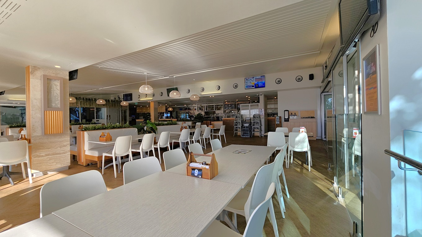 On the left, there are white tables and chairs near a wooden partition with potted plants on top. On the right, there are more white tables and chairs near floor-to-ceiling windows with a view of trees. In the background, there is a counter with bar stools, a menu board, and a display of drinks. The floor is wooden, and the ceiling has white panels with recessed lighting and hanging spherical lamps. On the left, there are white tables and chairs near a wooden partition with potted plants on top. On the right, there are more white tables and chairs near floor-to-ceiling windows with a view of trees. In the background, there is a counter with bar stools, a menu board, and a display of drinks. The floor is wooden, and the ceiling has white panels with recessed lighting and hanging spherical lamps.