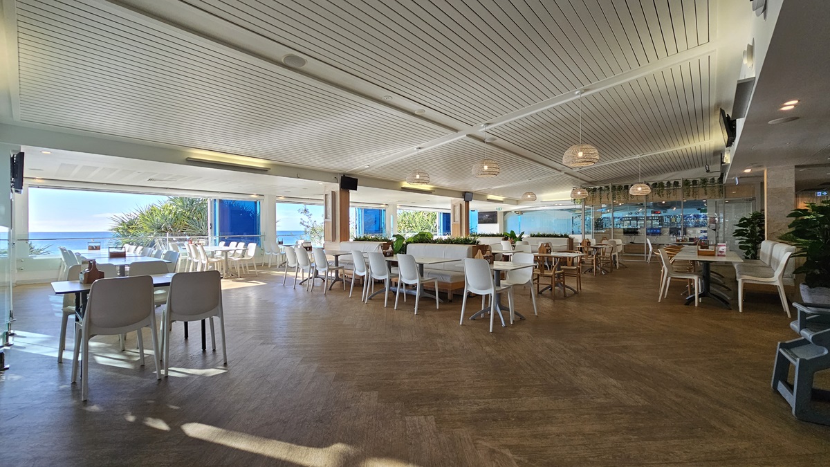 On the left, there are white tables and chairs near large windows with a view of the beach and trees. On the right, there is a bar area with stools, shelves of bottles, and more white tables and chairs. In the middle, there are additional tables and chairs, with a circular wooden partition featuring potted plants. The floor is wooden, and the ceiling has white panels with recessed lighting and hanging spherical lamps. On the left, there are white tables and chairs near large windows with a view of the beach and trees. On the right, there is a bar area with stools, shelves of bottles, and more white tables and chairs. In the middle, there are additional tables and chairs, with a circular wooden partition featuring potted plants. The floor is wooden, and the ceiling has white panels with recessed lighting and hanging spherical lamps.