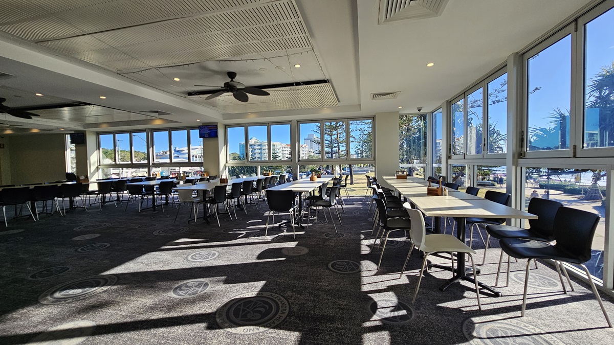 On the left, there are rows of black chairs and white tables near large windows. On the right, there are more white tables and black chairs near floor-to-ceiling windows with views of trees and buildings. In the middle, there are additional tables and chairs with sunlight streaming through the windows. The floor is carpeted with circular patterns, and the ceiling has white panels with recessed lighting and ceiling fans. On the left, there are rows of black chairs and white tables near large windows. On the right, there are more white tables and black chairs near floor-to-ceiling windows with views of trees and buildings. In the middle, there are additional tables and chairs with sunlight streaming through the windows. The floor is carpeted with circular patterns, and the ceiling has white panels with recessed lighting and ceiling fans.