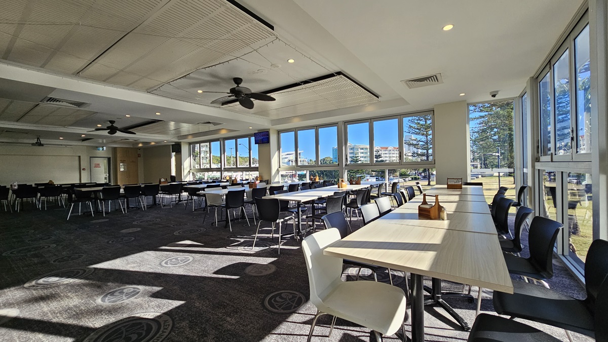On the left, there are rows of black chairs and white tables near large windows. On the right, there are more white tables and black chairs near floor-to-ceiling windows with views of trees and buildings. In the middle, there are additional tables and chairs with sunlight streaming through the windows. The floor is carpeted with circular patterns, and the ceiling has white panels with recessed lighting and ceiling fans. On the left, there are rows of black chairs and white tables near large windows. On the right, there are more white tables and black chairs near floor-to-ceiling windows with views of trees and buildings. In the middle, there are additional tables and chairs with sunlight streaming through the windows. The floor is carpeted with circular patterns, and the ceiling has white panels with recessed lighting and ceiling fans.