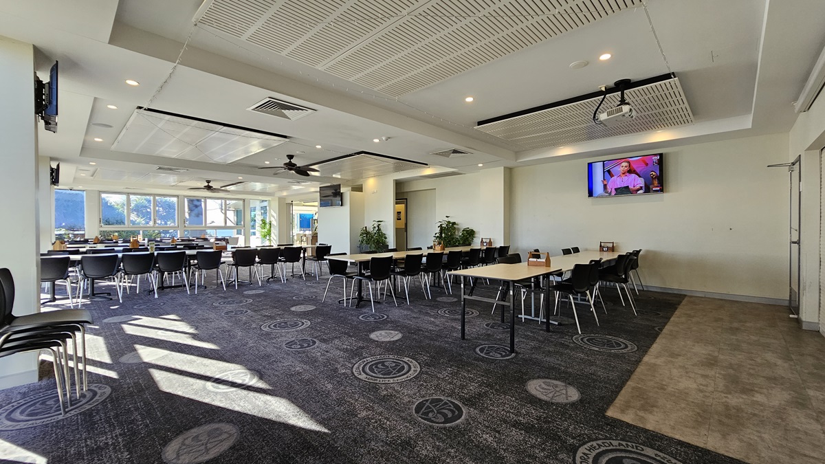 On the left, there are rows of black chairs and white tables near large windows with sunlight streaming in. On the right, there are more black chairs and white tables, with a television mounted on the wall displaying a programme. In the background, there is a bar area with stools and additional seating. The floor is carpeted with circular patterns, and the ceiling has white panels with recessed lighting, air conditioning vents, and ceiling fans. On the left, there are rows of black chairs and white tables near large windows with sunlight streaming in. On the right, there are more black chairs and white tables, with a television mounted on the wall displaying a programme. In the background, there is a bar area with stools and additional seating. The floor is carpeted with circular patterns, and the ceiling has white panels with recessed lighting, air conditioning vents, and ceiling fans.