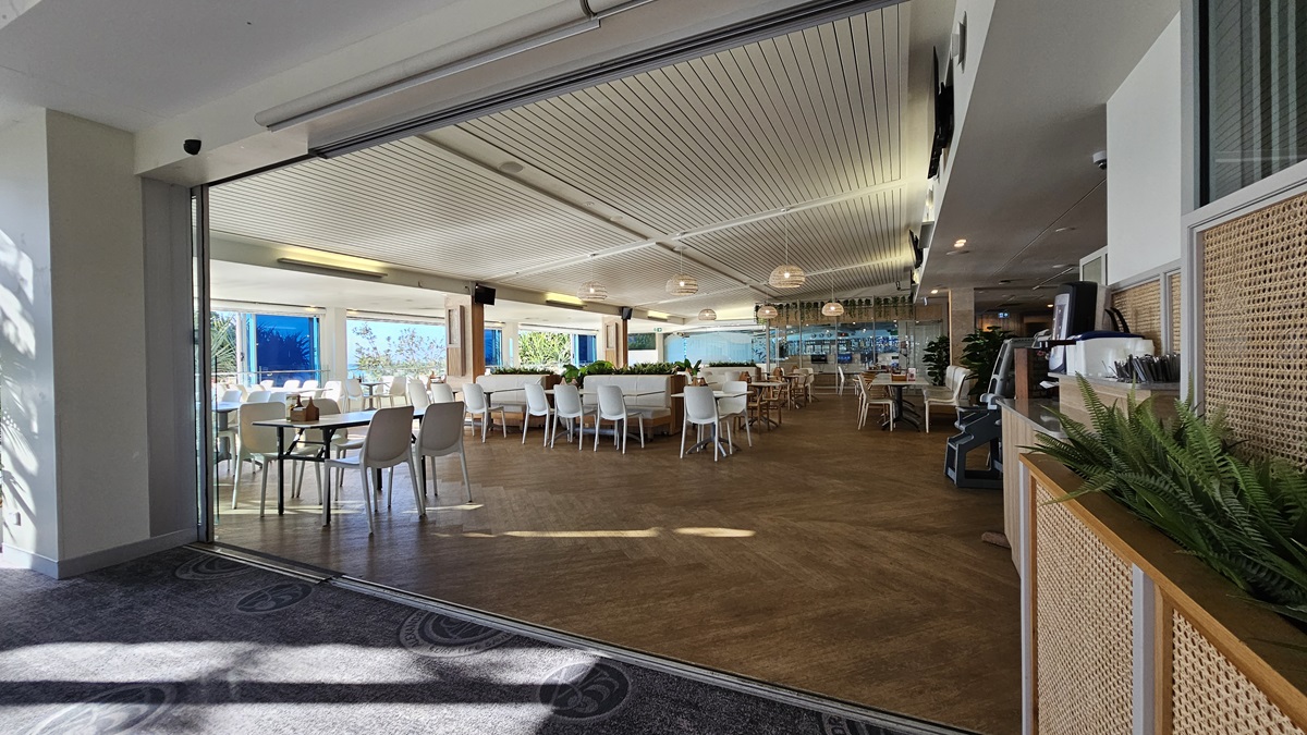 On the left, there are white tables and chairs near large windows with a view of trees and the sea. On the right, there is a wooden counter with potted plants and a coffee machine. In the middle, there are more white tables and chairs arranged in rows. The floor is a mix of wooden and carpeted areas, and the ceiling has white panels with recessed lighting and hanging spherical lamps. On the left, there are white tables and chairs near large windows with a view of trees and the sea. On the right, there is a wooden counter with potted plants and a coffee machine. In the middle, there are more white tables and chairs arranged in rows. The floor is a mix of wooden and carpeted areas, and the ceiling has white panels with recessed lighting and hanging spherical lamps.