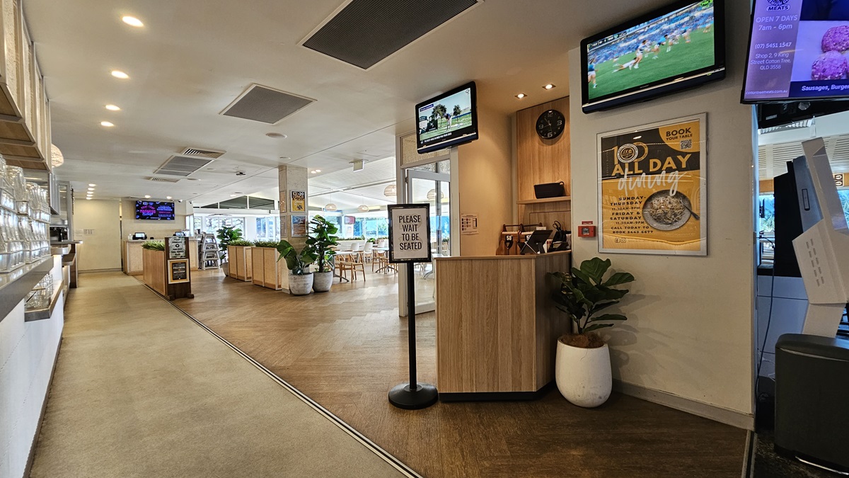 On the left, there is a long counter with a display case and a row of overhead lights. In the middle, there is a wooden reception desk with a sign that reads "Please Wait to be Seated," and a potted plant beside it. In the background, there are dining tables with chairs and another counter area. On the right, there are two wall-mounted screens, one showing a sports channel and the other displaying a menu. The floor is a mix of carpet and wood, and the ceiling has recessed lighting. On the left, there is a long counter with a display case and a row of overhead lights. In the middle, there is a wooden reception desk with a sign that reads "Please Wait to be Seated," and a potted plant beside it. In the background, there are dining tables with chairs and another counter area. On the right, there are two wall-mounted screens, one showing a sports channel and the other displaying a menu. The floor is a mix of carpet and wood, and the ceiling has recessed lighting.