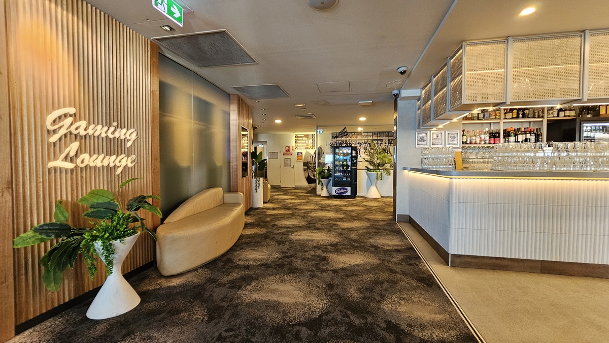 On the left, there is a wooden wall with a neon sign reading "Gaming Lounge" and a white sofa next to potted plants. In the middle, there is a hallway with a dark, patterned carpet leading to a vending machine and a door in the background. On the right, there is a long bar counter with stools, shelves stocked with bottles, and glasses hanging overhead. The ceiling has recessed lighting and air conditioning vents. On the left, there is a wooden wall with a neon sign reading "Gaming Lounge" and a white sofa next to potted plants. In the middle, there is a hallway with a dark, patterned carpet leading to a vending machine and a door in the background. On the right, there is a long bar counter with stools, shelves stocked with bottles, and glasses hanging overhead. The ceiling has recessed lighting and air conditioning vents.