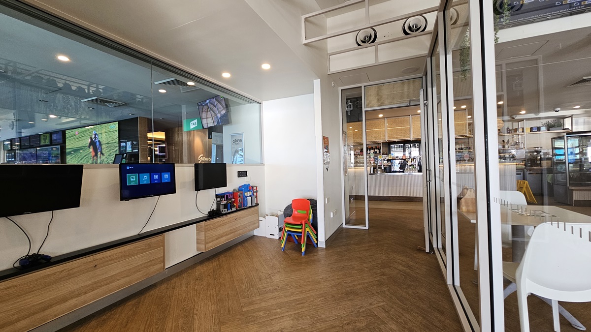 On the left, there are wall-mounted screens displaying various images and information, with wooden cabinets below them. In the middle, there is a small stack of colourful children's chairs and a red high chair. In the background, there is a bar area with stools, shelves stocked with bottles, and a counter. On the right, there are glass doors and windows leading to the bar area, with a white chair nearby. The floor is wooden, and the ceiling has recessed lighting and air conditioning vents. On the left, there are wall-mounted screens displaying various images and information, with wooden cabinets below them. In the middle, there is a small stack of colourful children's chairs and a red high chair. In the background, there is a bar area with stools, shelves stocked with bottles, and a counter. On the right, there are glass doors and windows leading to the bar area, with a white chair nearby. The floor is wooden, and the ceiling has recessed lighting and air conditioning vents.