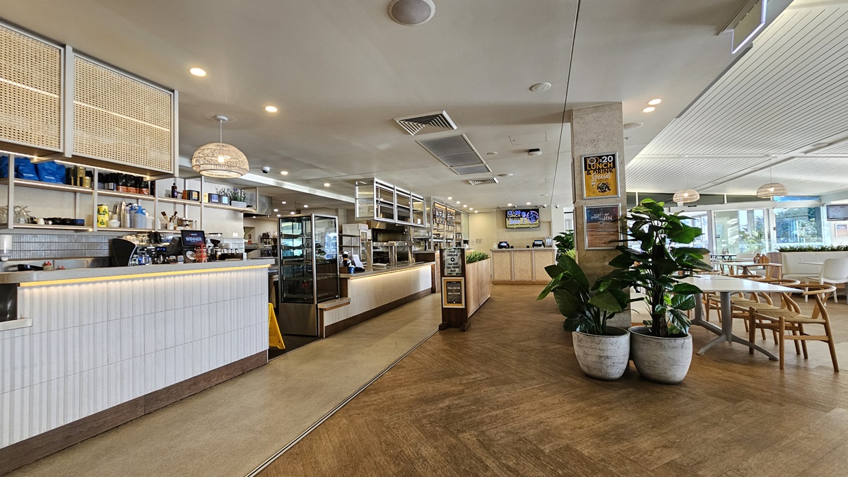 On the left, there is a long counter with a white front, illuminated from below, and a display case with pastries. Behind the counter are shelves with bottles and coffee machines. In the middle, there is a wooden partition with potted plants in front of it. In the background, there is another counter area with a digital screen above it. On the right, there are tables and chairs near large windows. The floor is wooden, and the ceiling has recessed lighting and air conditioning vents. On the left, there is a long counter with a white front, illuminated from below, and a display case with pastries. Behind the counter are shelves with bottles and coffee machines. In the middle, there is a wooden partition with potted plants in front of it. In the background, there is another counter area with a digital screen above it. On the right, there are tables and chairs near large windows. The floor is wooden, and the ceiling has recessed lighting and air conditioning vents.