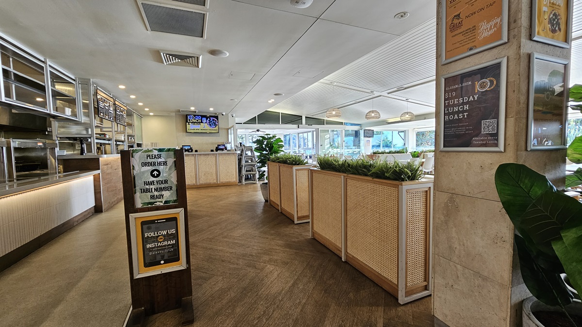 On the left, there is a long counter with a white front, a "Please Order Here" sign, and shelves with various items above. In the middle, there is a wooden partition with potted plants on top, leading to a dining area with tables and chairs near large windows. In the background, there is a digital screen and more seating. On the right, there are posters on the wall, a potted plant, and a sign about a lunch roast. The floor is wooden, and the ceiling has recessed lighting and air con vents. On the left, there is a long counter with a white front, a "Please Order Here" sign, and shelves with various items above. In the middle, there is a wooden partition with potted plants on top, leading to a dining area with tables and chairs near large windows. In the background, there is a digital screen and more seating. On the right, there are posters on the wall, a potted plant, and a sign about a lunch roast. The floor is wooden, and the ceiling has recessed lighting and air con vents.