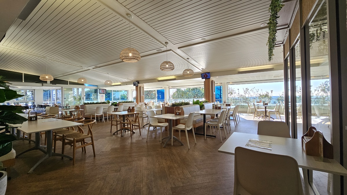 On the left, there are several tables and chairs near large windows with a view of the sea. In the middle, there are more tables and chairs, with a wooden partition featuring potted plants on top. In the background, there are additional seating areas and large windows showcasing the sea view. On the right, there are more tables and chairs near floor-to-ceiling windows. The floor is wooden, and the ceiling has white panels with recessed lighting and hanging wicker lamps. On the left, there are several tables and chairs near large windows with a view of the sea. In the middle, there are more tables and chairs, with a wooden partition featuring potted plants on top. In the background, there are additional seating areas and large windows showcasing the sea view. On the right, there are more tables and chairs near floor-to-ceiling windows. The floor is wooden, and the ceiling has white panels with recessed lighting and hanging wicker lamps.