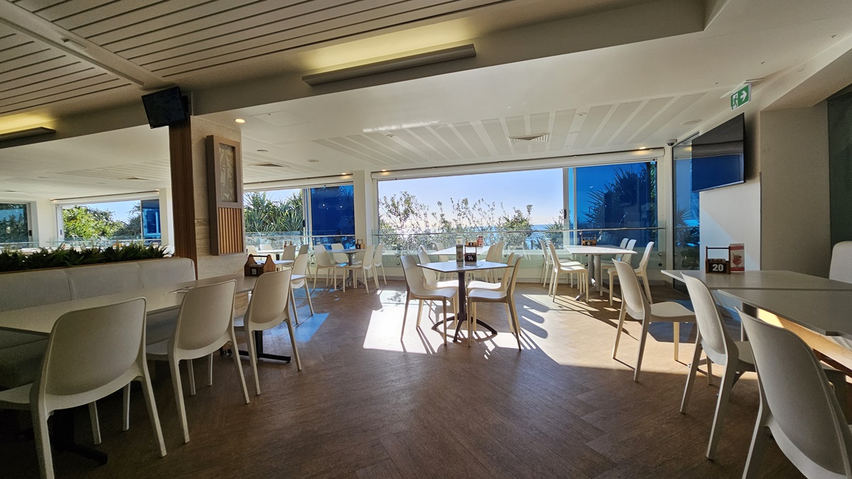 On the left, there are white tables and chairs arranged near a wooden partition with potted plants on top. In the middle, there are more white tables and chairs, with sunlight streaming through large windows. In the background, there are additional seating areas and large windows showcasing a view of trees and foliage. On the right, there are more tables and chairs near floor-to-ceiling windows with blue blinds. The floor is wooden, and the ceiling has white panels with recessed lighting. On the left, there are white tables and chairs arranged near a wooden partition with potted plants on top. In the middle, there are more white tables and chairs, with sunlight streaming through large windows. In the background, there are additional seating areas and large windows showcasing a view of trees and foliage. On the right, there are more tables and chairs near floor-to-ceiling windows with blue blinds. The floor is wooden, and the ceiling has white panels with recessed lighting.