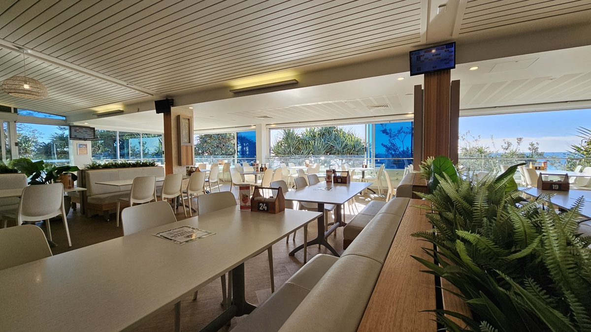 On the left, there are white tables and chairs near large windows with a view of trees and foliage. In the middle, there are more tables and chairs, with a wooden partition featuring potted plants on top. In the background, there are additional seating areas and large windows with blue blinds. On the right, there are more tables and chairs near floor-to-ceiling windows. The floor is wooden, and the ceiling has white panels with recessed lighting and a mounted TV screen. On the left, there are white tables and chairs near large windows with a view of trees and foliage. In the middle, there are more tables and chairs, with a wooden partition featuring potted plants on top. In the background, there are additional seating areas and large windows with blue blinds. On the right, there are more tables and chairs near floor-to-ceiling windows. The floor is wooden, and the ceiling has white panels with recessed lighting and a mounted TV screen.