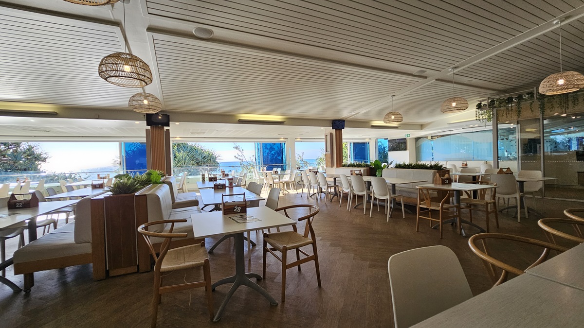On the left, there are white tables and chairs near large windows with a view of the sea and trees. In the middle, there are more tables and chairs, with a wooden partition, potted plants on top. In the background, there are additional seating areas and large windows with blue blinds. On the right, there are more tables and chairs near floor-to-ceiling windows, with a section of the room separated by glass walls. The floor is wooden, and the ceiling has white panels with recessed lighting. On the left, there are white tables and chairs near large windows with a view of the sea and trees. In the middle, there are more tables and chairs, with a wooden partition, potted plants on top. In the background, there are additional seating areas and large windows with blue blinds. On the right, there are more tables and chairs near floor-to-ceiling windows, with a section of the room separated by glass walls. The floor is wooden, and the ceiling has white panels with recessed lighting.