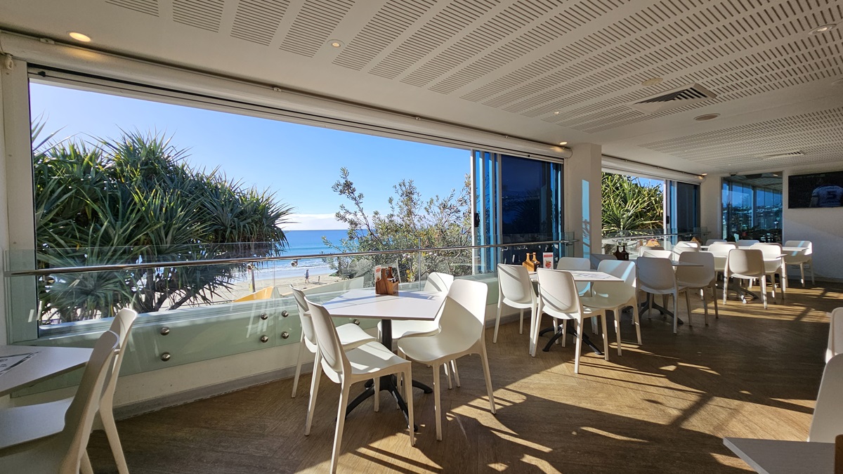 On the left, there are white tables and chairs near large windows with a view of the beach and trees. On the right, there are more white tables and chairs near floor-to-ceiling windows, with blue blinds partially drawn. In the middle, there are additional tables and chairs, with sunlight streaming through the windows. The floor is wooden, and the ceiling has white panels with recessed lighting and air conditioning vents. On the left, there are white tables and chairs near large windows with a view of the beach and trees. On the right, there are more white tables and chairs near floor-to-ceiling windows, with blue blinds partially drawn. In the middle, there are additional tables and chairs, with sunlight streaming through the windows. The floor is wooden, and the ceiling has white panels with recessed lighting and air conditioning vents.