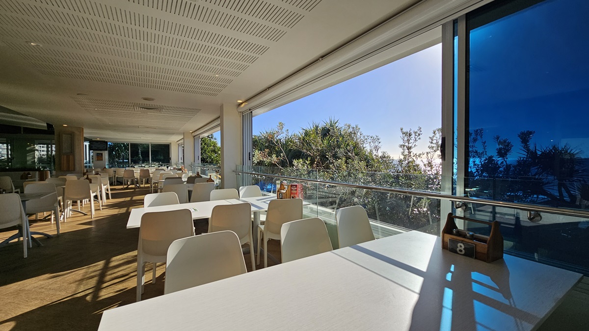 On the left, there are white tables and chairs near large windows with a view of the beach and trees. On the right, there are more white tables and chairs near floor-to-ceiling windows with blue-tinted glass. In the middle, there are additional tables and chairs, with a wooden partition featuring potted plants on top. The floor is wooden, and the ceiling has white panels with recessed lighting, air conditioning vents, and hanging wicker lamps. On the left, there are white tables and chairs near large windows with a view of the beach and trees. On the right, there are more white tables and chairs near floor-to-ceiling windows with blue-tinted glass. In the middle, there are additional tables and chairs, with a wooden partition featuring potted plants on top. The floor is wooden, and the ceiling has white panels with recessed lighting, air conditioning vents, and hanging wicker lamps.