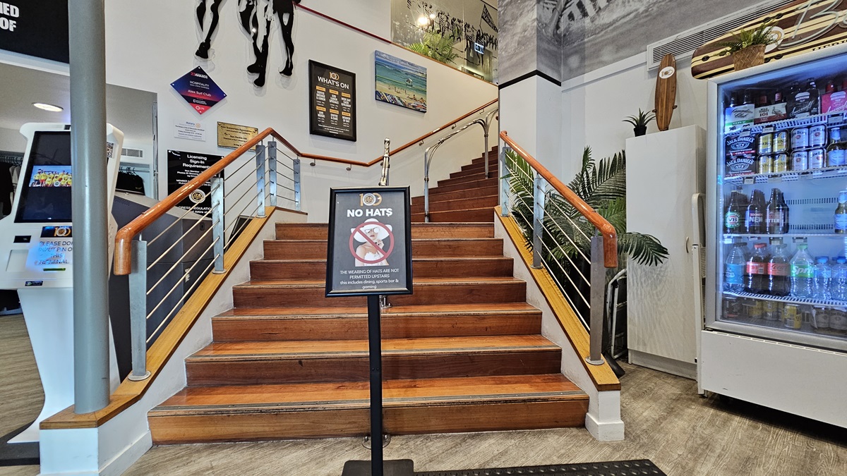 On the left, there is a white kiosk with a touchscreen and a wall with various signs. In the middle, there is a wooden staircase with metal railings leading up to a landing, with a "No Hats" sign at the base. In the background, there are framed pictures and a plant on the landing. On the right, there is a large refrigerator stocked with drinks and a tall potted plant beside it. The floor is light-coloured wood, and the ceiling has white panels with recessed lighting. On the left, there is a white kiosk with a touchscreen and a wall with various signs. In the middle, there is a wooden staircase with metal railings leading up to a landing, with a "No Hats" sign at the base. In the background, there are framed pictures and a plant on the landing. On the right, there is a large refrigerator stocked with drinks and a tall potted plant beside it. The floor is light-coloured wood, and the ceiling has white panels with recessed lighting.