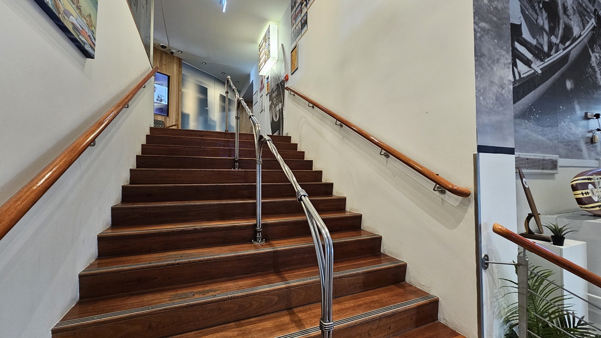 On the left, there is a wooden handrail along the wall with framed pictures above it. In the middle, a wooden staircase with metal railings leads up to a landing, where a door and more framed pictures are visible. On the right, there is another wooden handrail, a large black-and-white mural on the wall, and a potted plant at the base of the stairs. The floor is wooden, and the ceiling has recessed lighting and a small light fixture near the top of the stairs. On the left, there is a wooden handrail along the wall with framed pictures above it. In the middle, a wooden staircase with metal railings leads up to a landing, where a door and more framed pictures are visible. On the right, there is another wooden handrail, a large black-and-white mural on the wall, and a potted plant at the base of the stairs. The floor is wooden, and the ceiling has recessed lighting and a small light fixture near the top of the stairs.